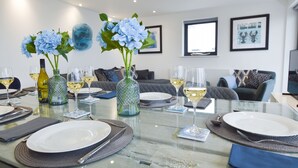 Un coin repas avec une table en verre mise pour un repas, agrémentée d’assiettes blanches, de couverts et de verres à vin. On y trouve deux vases décoratifs avec des fleurs bleues et une bouteille de vin blanc. La pièce présente un design moderne, avec un canapé gris et un tableau encadré au mur.