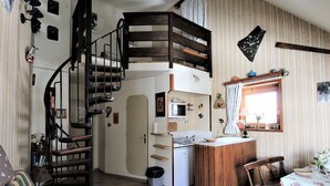 Cocina compacta con una isla central, zona de comedor con mesa y sillas, y una escalera de caracol que lleva a un nivel superior.