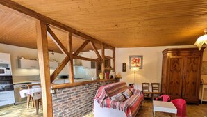 Ein gemütlicher Wohnbereich mit Holzbalkendecke, einer Ziegelwand, einem Sofa, einem Esstisch und einer Küche im Hintergrund.