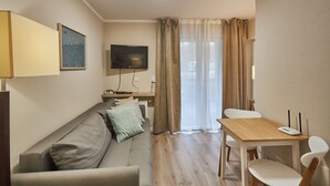 A modern living room with a sofa, a dining table with chairs, a TV mounted on the wall, and a window with curtains. - Apartamenty Swinoujscie - Bursztyn (Swinoujscie)