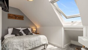 Une chambre avec un plafond en pente, un puits de lumière, un lit recouvert d’une couverture grise, une table de chevet avec une lampe et une œuvre d’art accrochée au mur.