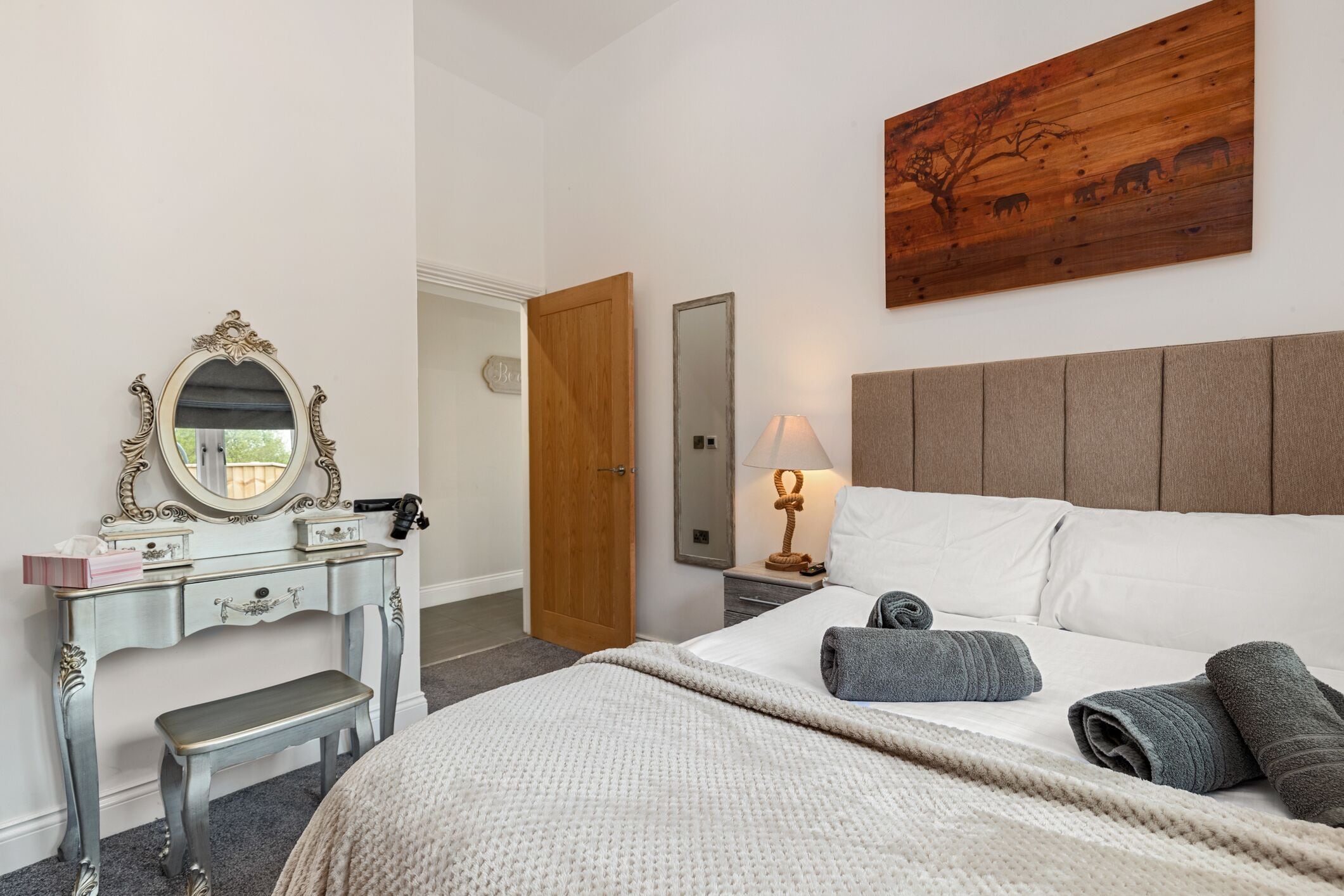 Een slaapkamer met een bed, een kaptafel met spiegel, een houten hoofdeinde en een kunstwerk aan de muur.