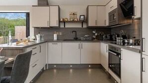 A modern kitchen with white cabinets, a built-in oven, and a microwave. There is a wooden shelf above the sink with various items. - Cedar Cottage (Tenby)