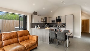 A modern kitchen with a central island, dining area with chairs, and a leather sofa. - Cedar Cottage (Tenby)