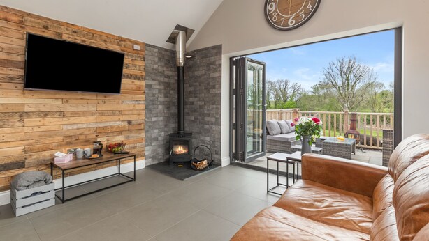 A modern living room with a fireplace, a TV mounted on a wooden wall, and a large window offering a view of the outdoors. - Cedar Cottage (Tenby)