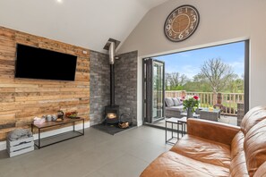 A modern living room with a fireplace, a TV mounted on a wooden wall, and a large window offering a view of the outdoors. - Cedar Cottage (Tenby)