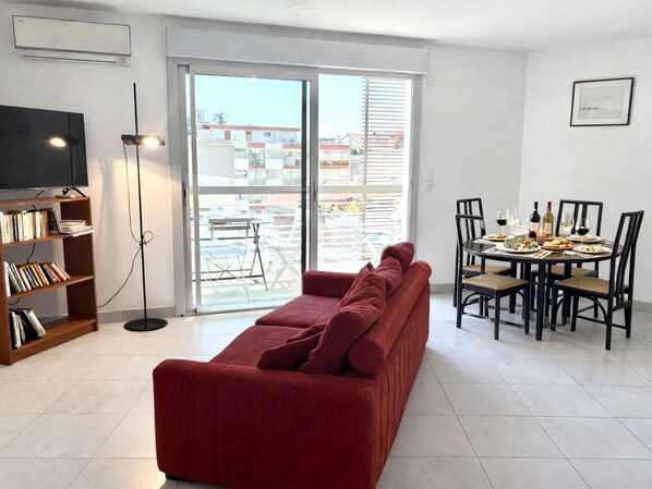 A living room with a red sofa, a dining table with chairs, a bookshelf, and a TV.