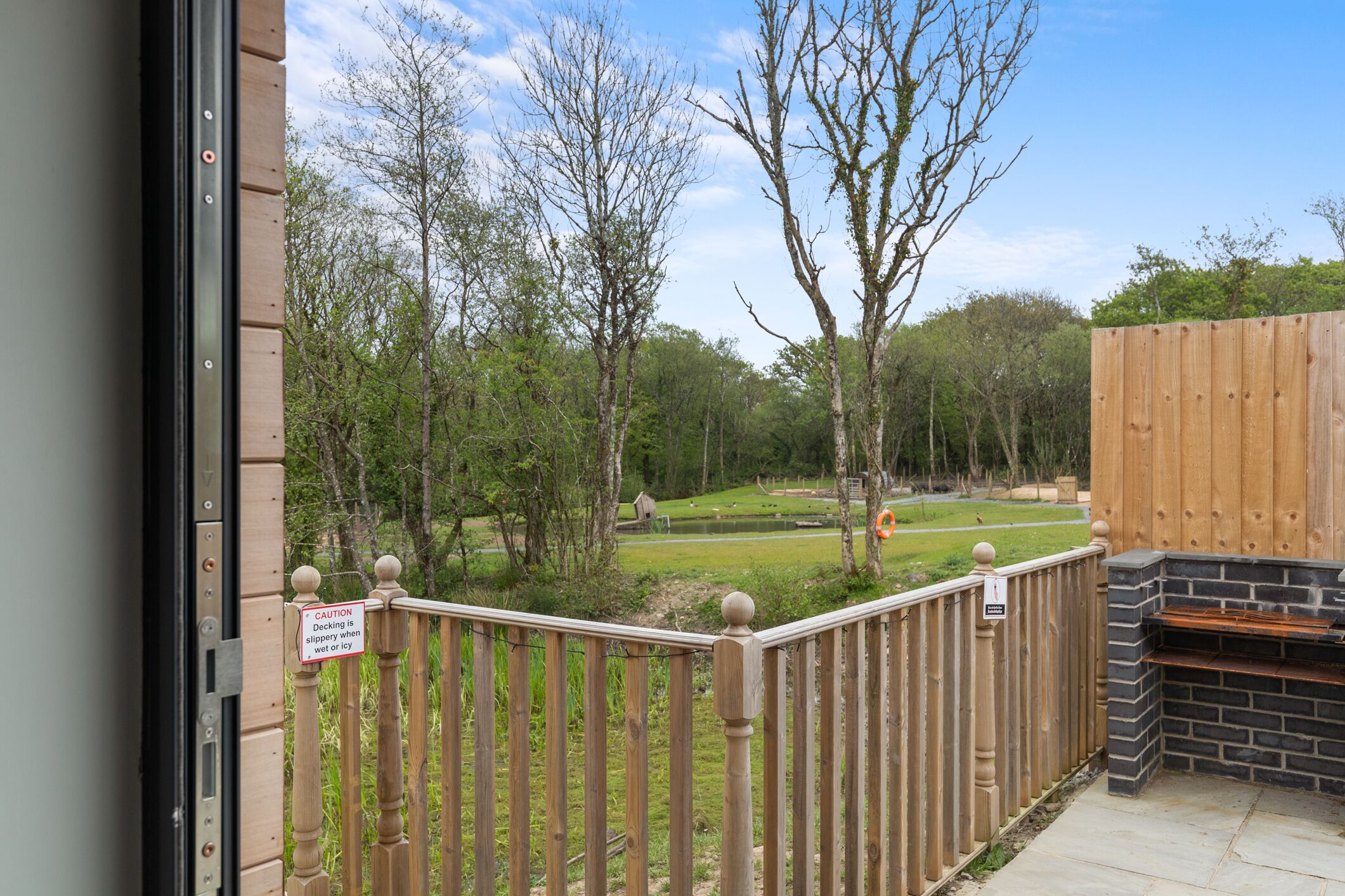 A wooden deck with a caution sign, a barbecue area, and a view of a park with trees and a pond.