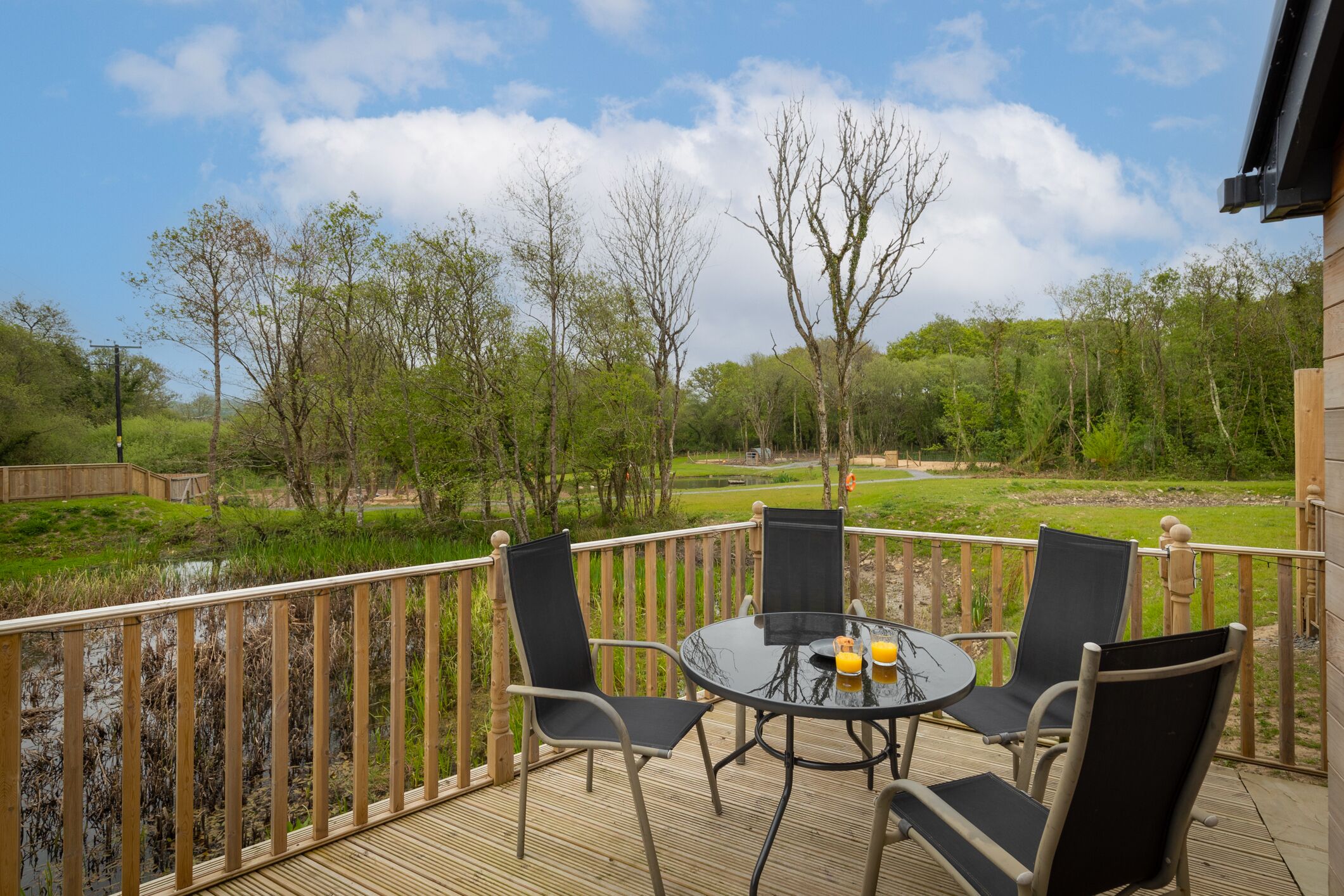 A wooden deck with a round table and four chairs, overlooking a grassy area with trees and a body of water.