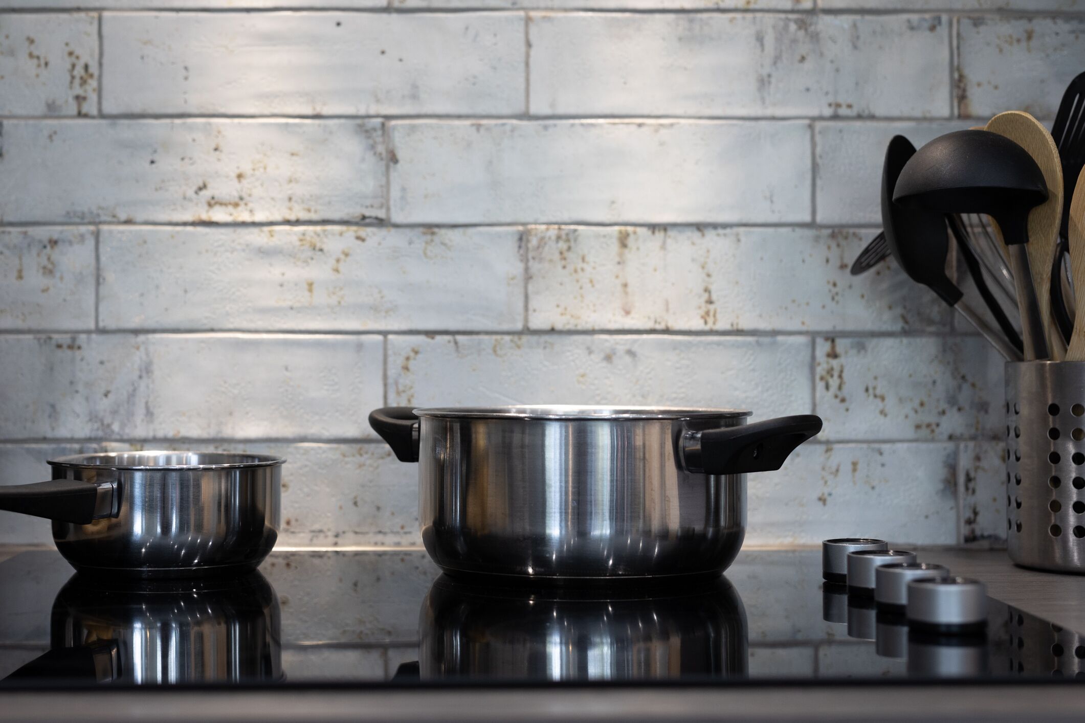 A kitchen with stainless steel pots on a stove, a brick wall, and a utensil holder with wooden and metal utensils.