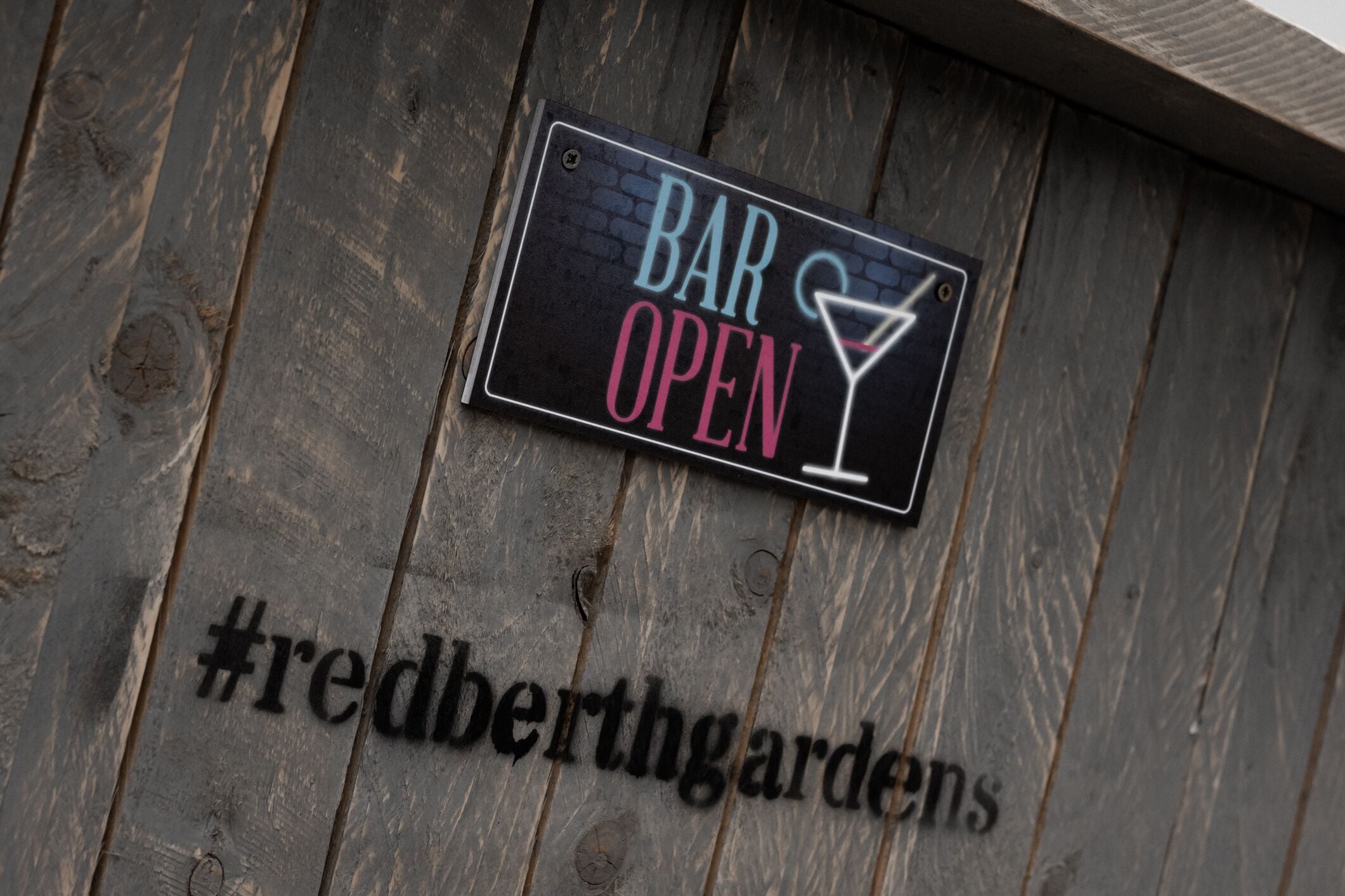 Un mur en bois sur lequel est affiché un panneau indiquant « BAR OUVERT » et le mot-dièse « #redberthgardens ».