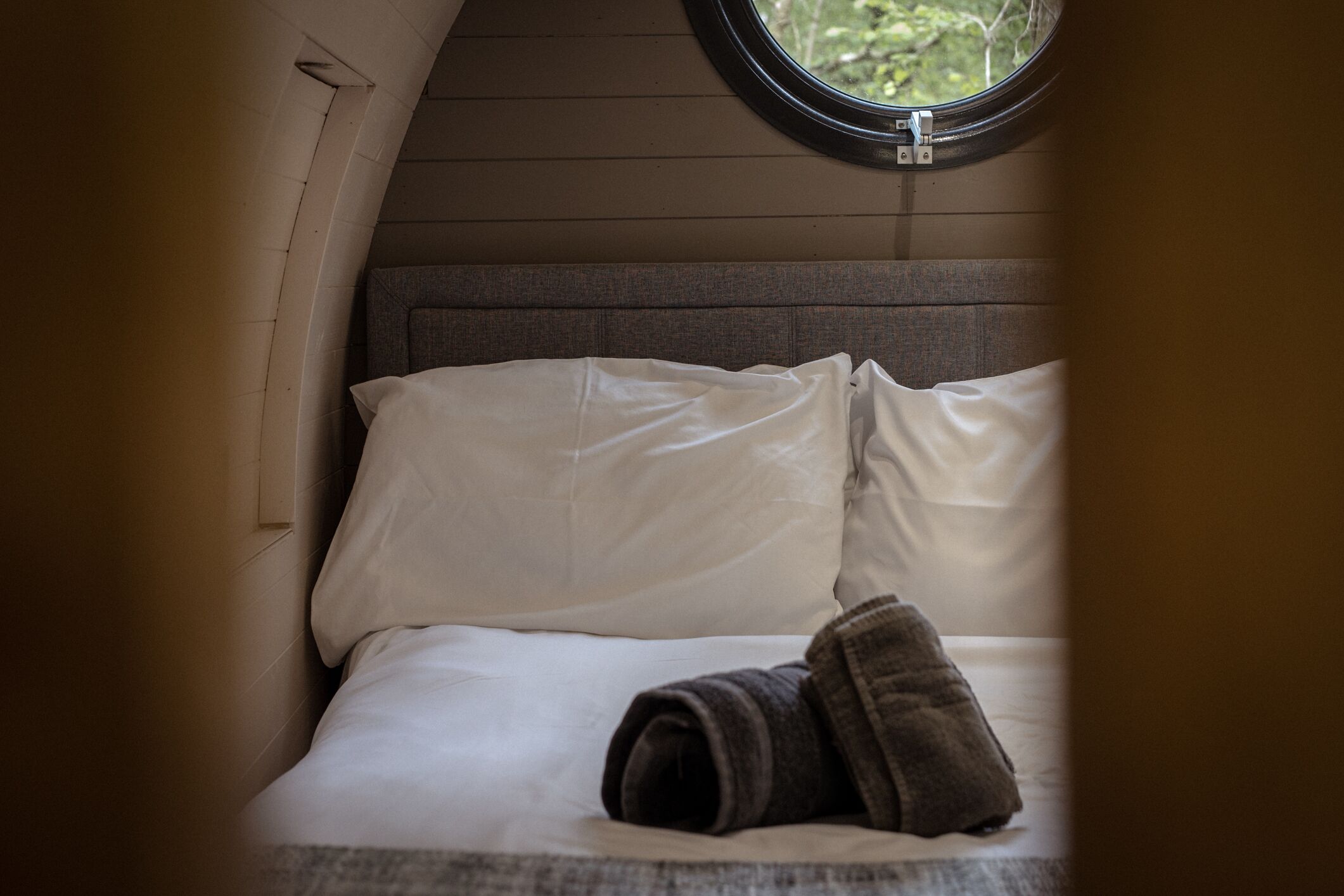 A bed with white linens and a rolled-up towel, a window with a view of greenery, and a wooden headboard.