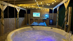 A hot tub with a television screen in the background, surrounded by wooden walls and string lights. - Calon (Tenby)