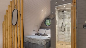 A bedroom with a sloped ceiling, a bed with a grey mattress and pillows, a wooden partition, a mirror, and a shower with a white tiled wall. - Calon (Tenby)