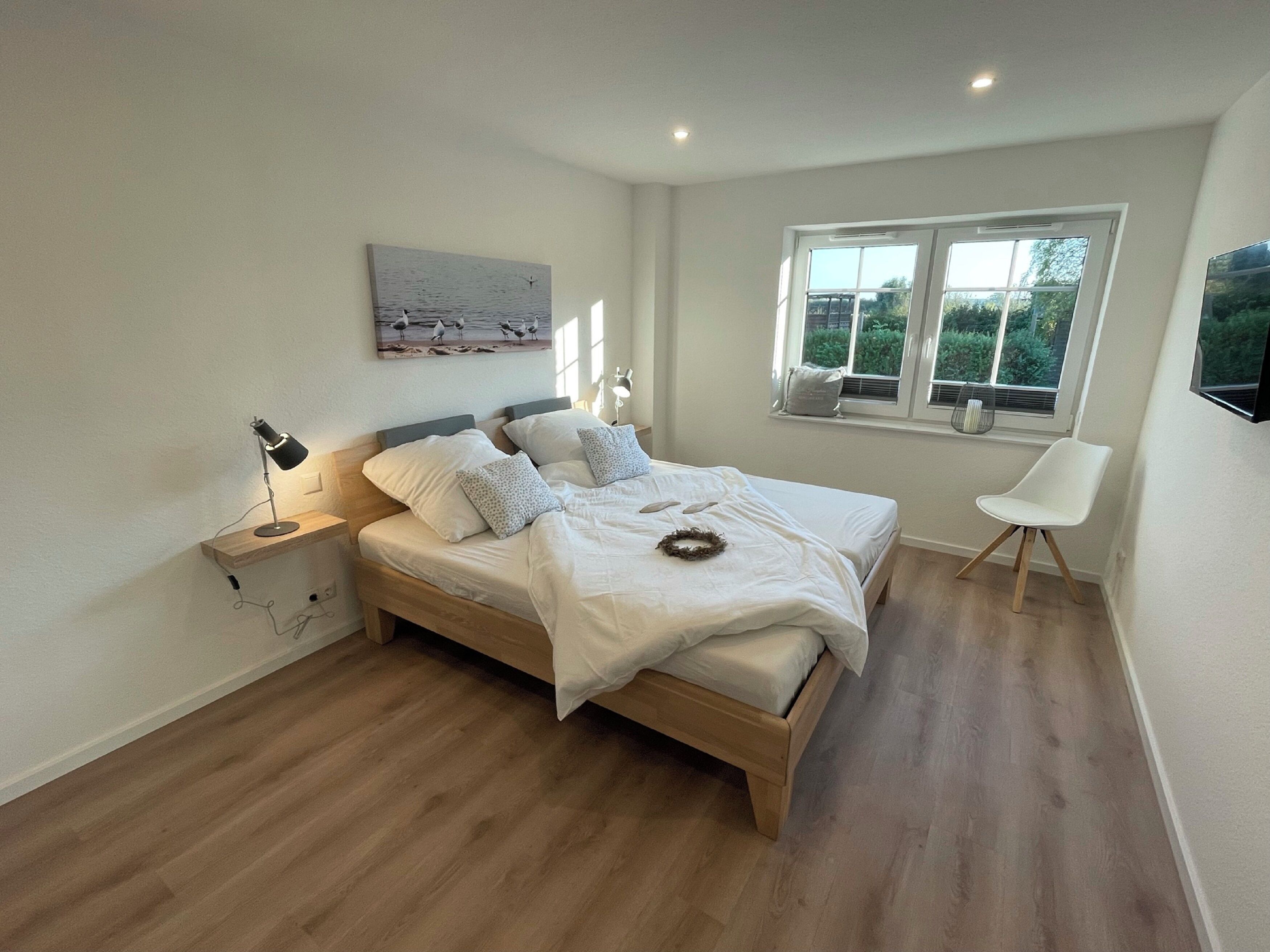 Un dormitorio con una cama de madera, una lámpara de noche, una ventana con vistas a zonas verdes y una silla blanca.