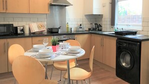A modern kitchen with wooden cabinets, a stainless steel range hood, a built-in washing machine, and a dining area with a round table and chairs. - Central Wigan Welcoming Townhouse Sleeps up to 6 (Wigan)