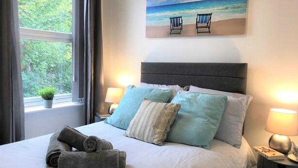 A neatly made bed with pillows and towels, a painting of beach chairs on the wall, and a window with curtains. - Central Wigan Welcoming Townhouse Sleeps up to 6 (Wigan)