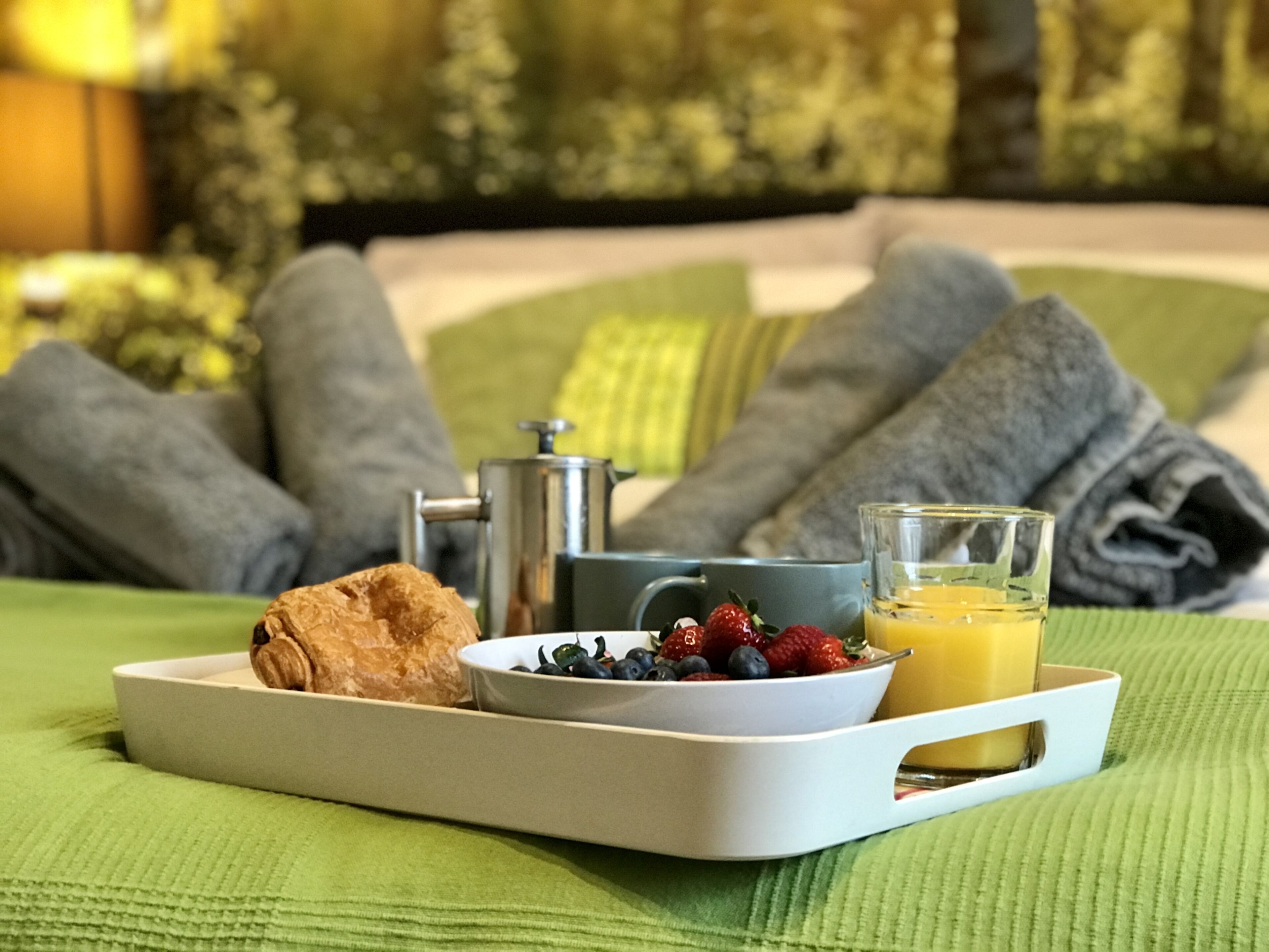 Ett frukostbricka med croissanter, bär och apelsinjuice, placerat på en säng med grönt sänglinne och grå kuddar.