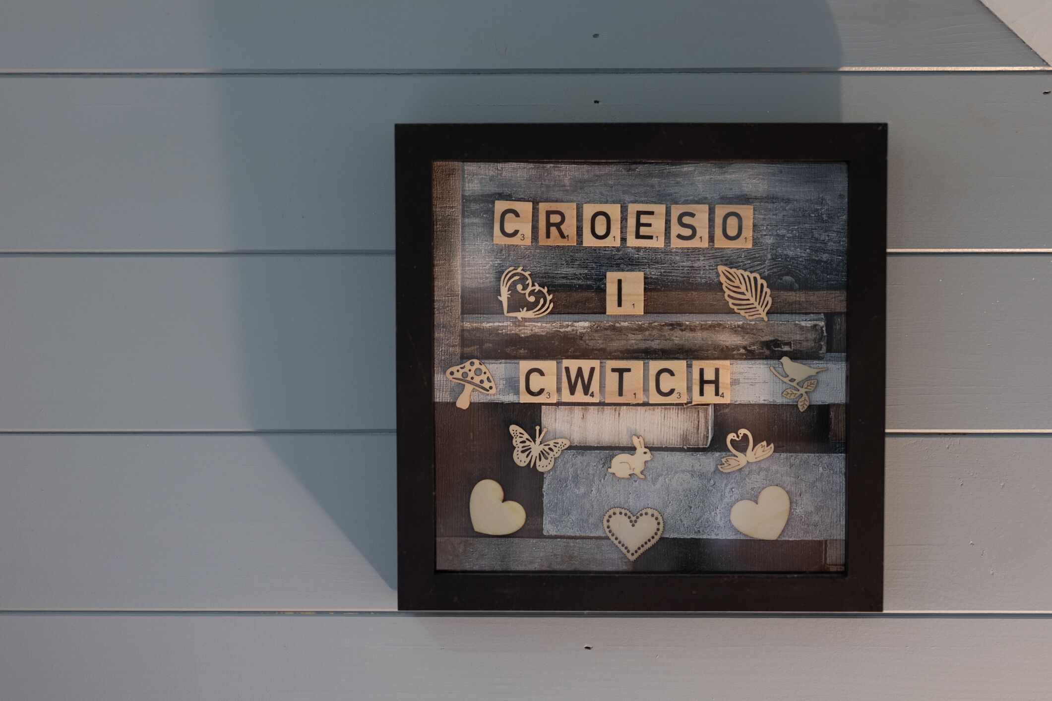 A framed artwork with the word 'CROESO' and 'CWTCH' on a wooden background, surrounded by decorative elements like leaves, a mushroom, hearts, and butterflies.