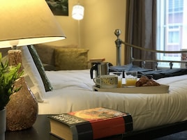 Una cama bien tendida con un libro, una bandeja con cruasanes, jugo y una cafetera, y una lámpara en una mesita de noche.