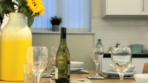 Une table à manger dressée avec de l’assiette blanche, des bols et des verres à vin, une bouteille de vin et un vase rempli de fleurs jaunes.