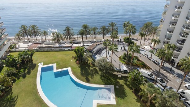 A swimming pool with a curved edge, surrounded by palm trees and a view of the ocean. - Bright Apartment with Front Sea Views (Marbella)