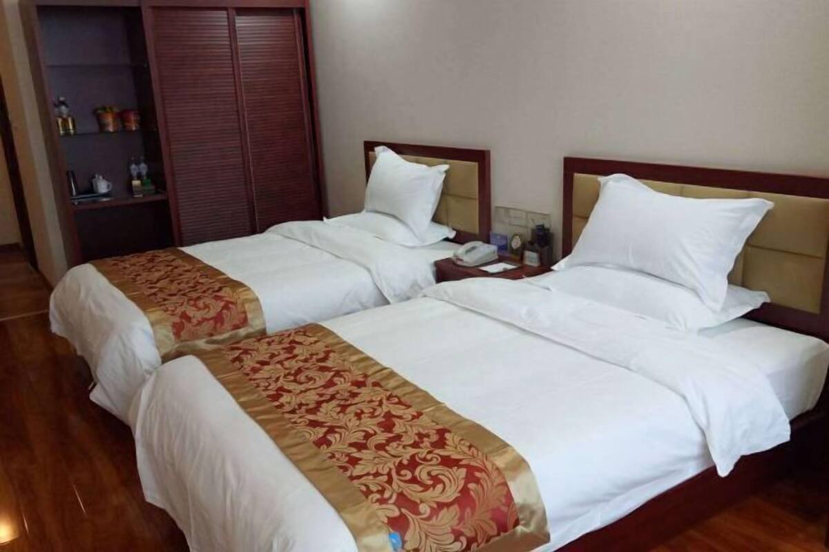 Une chambre d’hôtel avec deux lits, chacun agrémenté de draps blancs et d’une bordure décorative rouge et or. On y trouve une armoire en bois et une petite table de chevet avec une lampe et un téléphone.