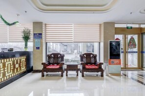 A hotel lobby with a reception desk, two wooden chairs, a potted plant, and a large window.