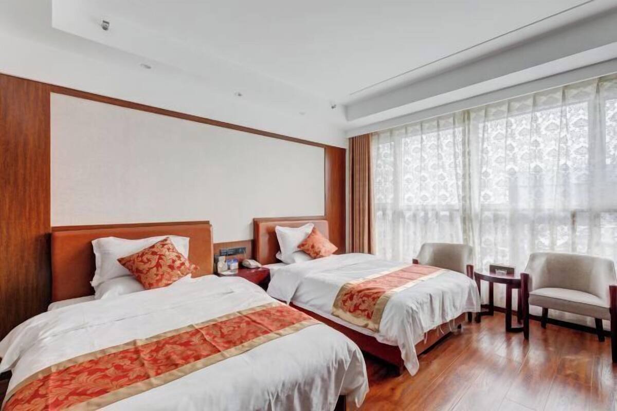Une chambre d’hôtel avec deux lits, une tête de lit en bois, une petite table, une chaise et une grande fenêtre avec des rideaux légers.