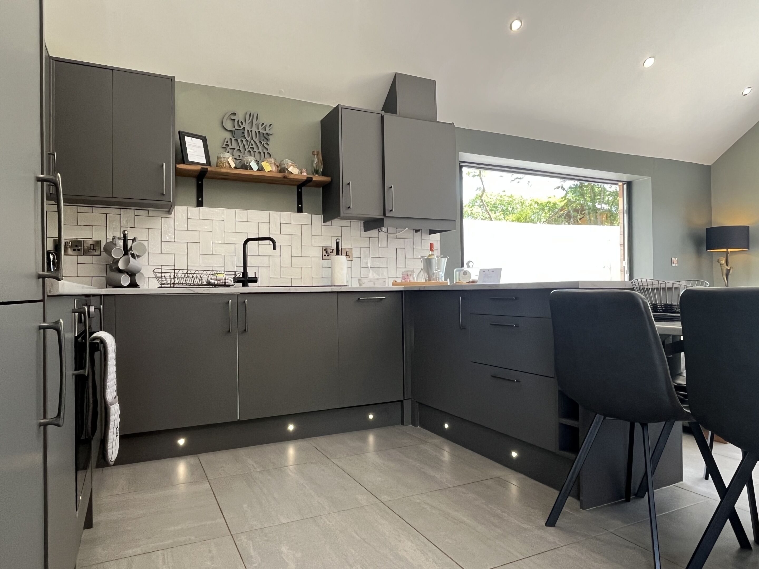 A modern kitchen with grey cabinets, a white tiled backsplash, and a window providing natural light.