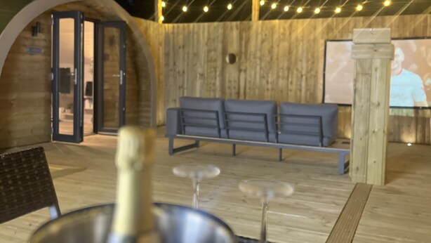 Un espacio al aire libre con piso de madera, un banco y una mesa con una botella y vasos.