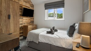 A bedroom with a wooden wall, a bed with a grey blanket, a TV mounted on the wall, a window with a view of trees, and a lamp on the bedside table.