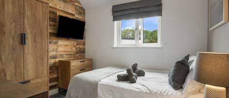Un dormitorio con pared de madera, una cama con manta gris, un televisor montado en la pared, una ventana con vista a árboles y una lámpara en la mesita de noche.