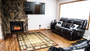 A living room with a stone fireplace, a black leather sofa, and a flat-screen TV mounted on the wall. - Central Home: Yard with Mountain Views, 3bdrm (Anchorage)