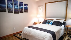 A neatly arranged hotel room with a large bed, a nightstand with a lamp, a window with blinds, and four framed pictures on the wall. - Central Home: Yard with Mountain Views, 3bdrm (Anchorage)