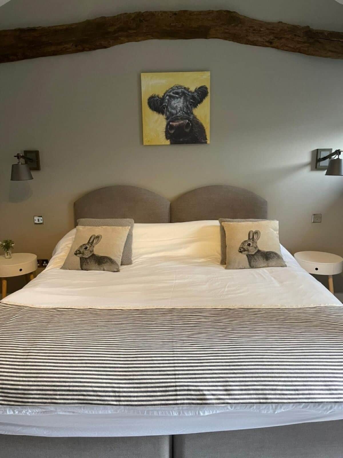 A bedroom with a bed, two pillows with rabbit designs, a painting of a dog, and a wooden beam ceiling.