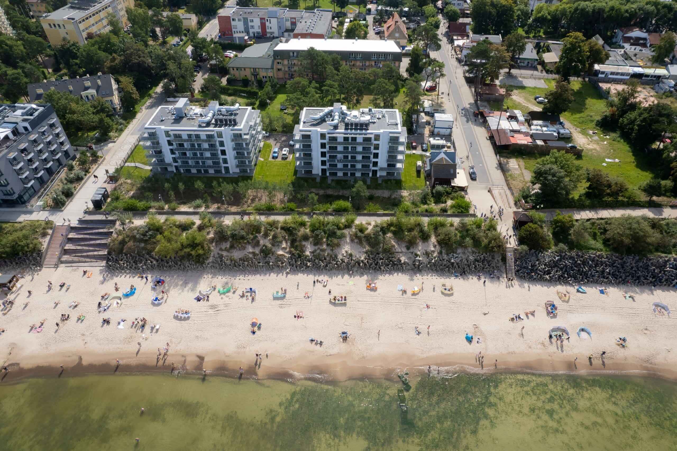 An aerial view of a beach with people and boats, surrounded by residential buildings and greenery.
