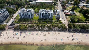 An aerial view of a beach with people and boats, surrounded by residential buildings and greenery.