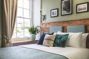 Un dormitorio con una cama grande, cabecera de madera y una ventana con cortinas. Hay cuadros enmarcados en la pared y una planta en maceta en el alféizar.