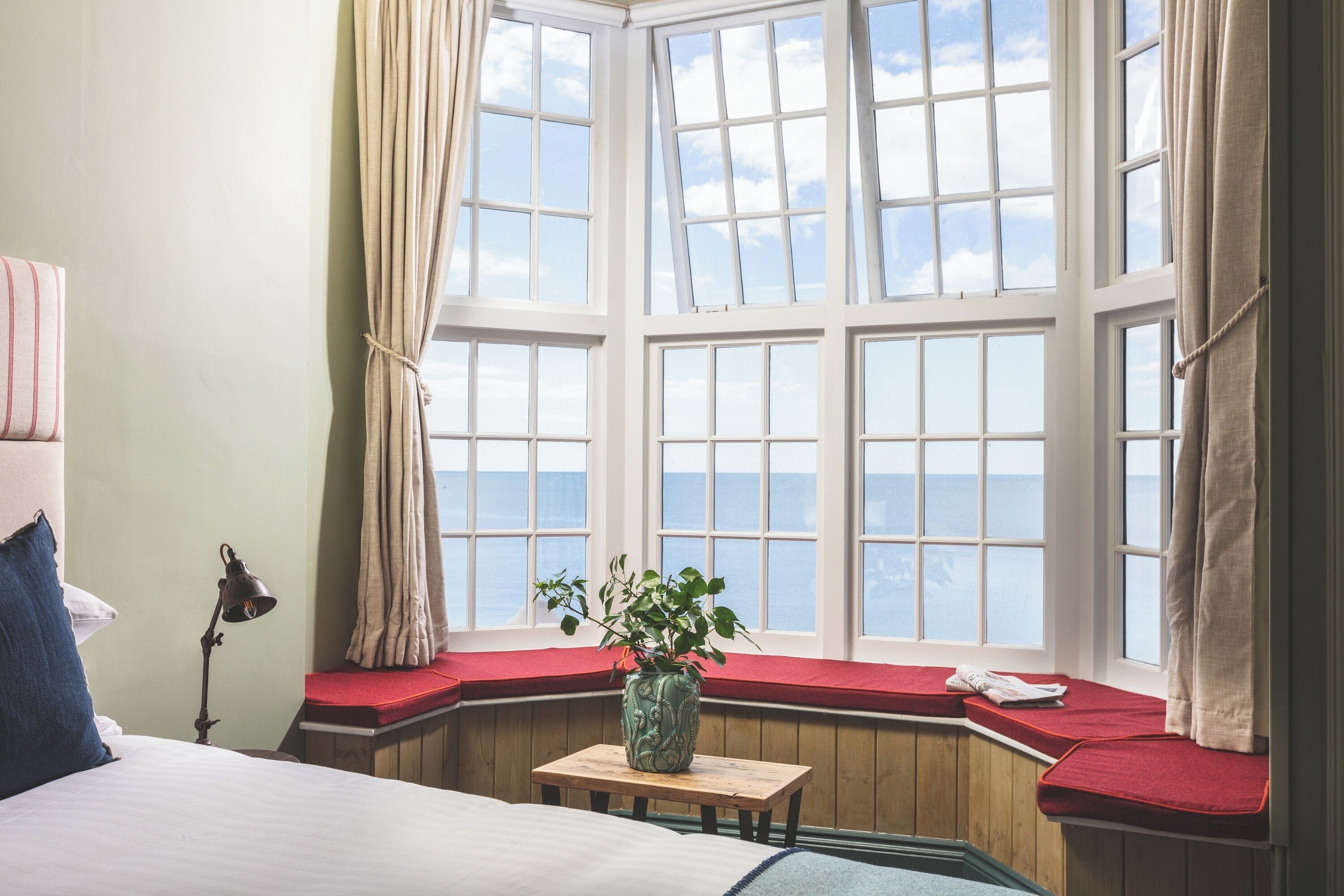 A room with a large bay window, a bed, a bedside lamp, a small table with a plant, and a red bench.