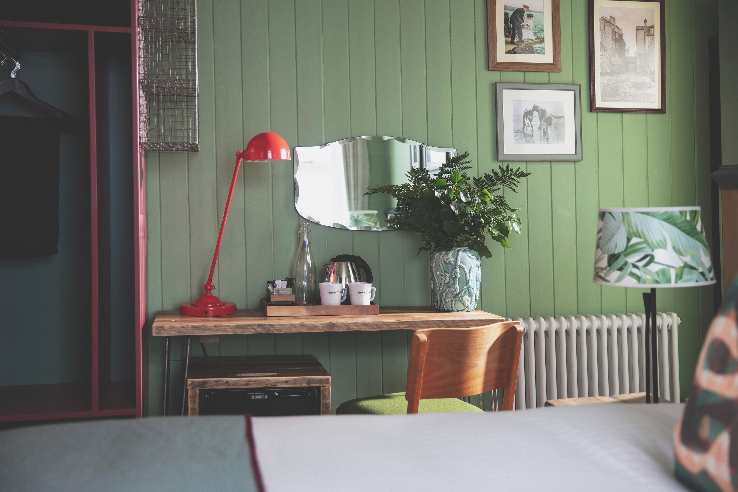 A room with a wooden desk, a red lamp, a mirror, a radiator, and framed pictures on the wall.