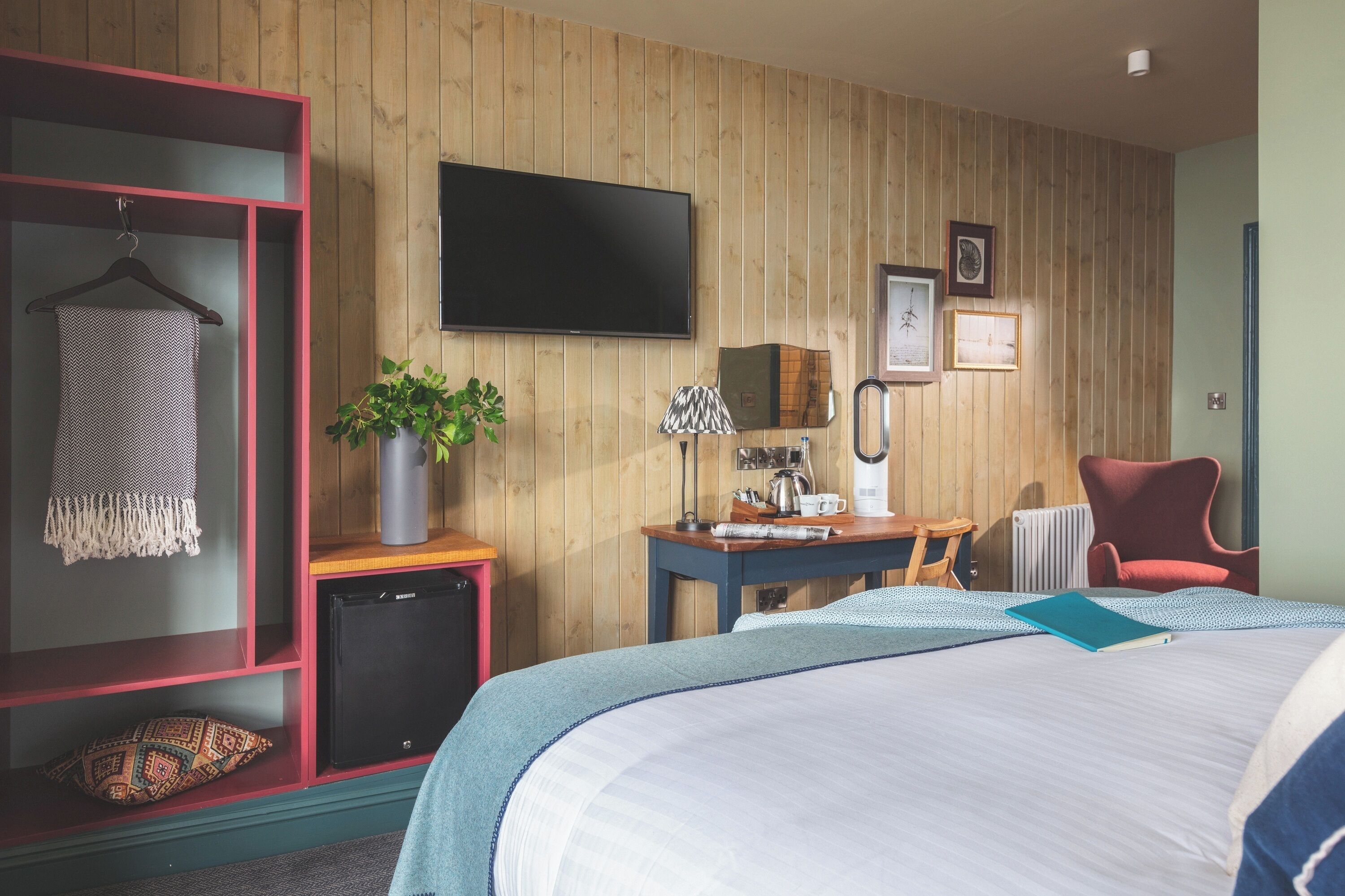 Quarto de hotel com cama, escrivaninha com luminária, televisão, cadeira vermelha e prateleira na parede com toalha e planta.