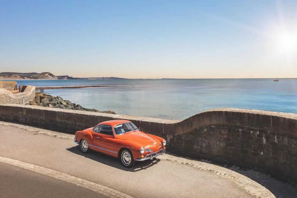 A vintage orange car parked on a coastal road with a clear view of the sea and distant hills.