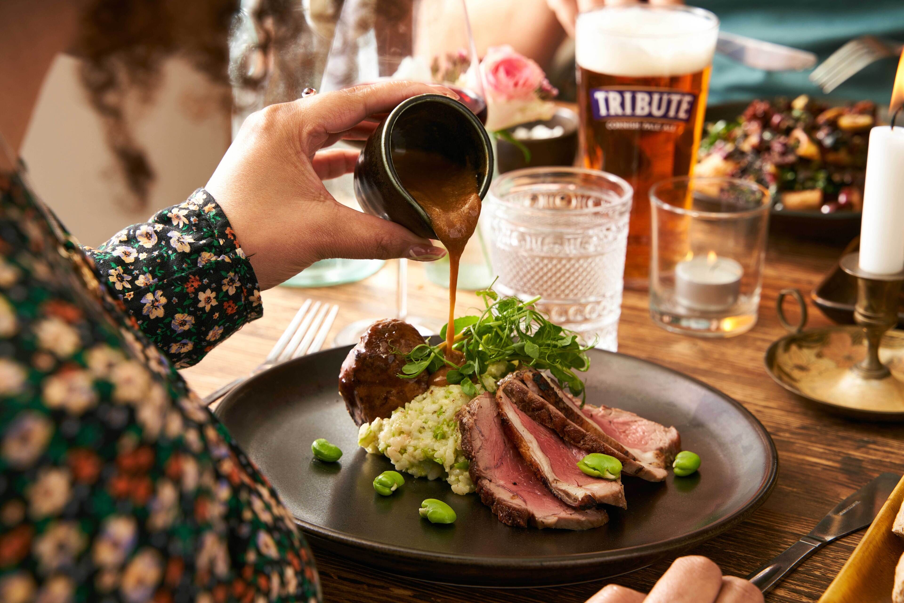 Una persona sirviendo salsa sobre un plato de comida, que incluye carne y verduras, con una copa de cerveza y una vela al fondo.