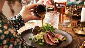 Una persona sirviendo salsa sobre un plato de comida, que incluye carne y verduras, con una copa de cerveza y una vela al fondo.