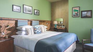 Habitación de hotel ordenada con cabecera de madera, una cama grande con colcha azul, una mesita de noche de madera con lámpara y una silla.