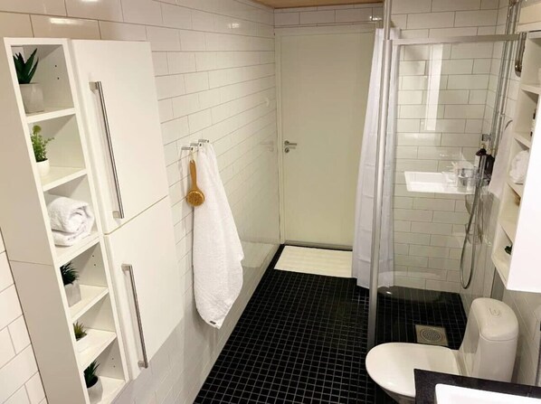 A modern bathroom with white tiled walls, a black tiled shower floor, a white toilet, and a glass-enclosed shower. - Superb 1BR home with sauna and terrace (Helsinki)