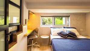 A compact bedroom with a desk, a bed with pillows, a window, and a small round stool.