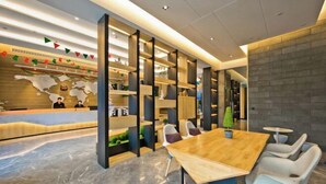 Un área de recepción moderna con un mostrador de madera, una mesa grande de madera y sillas blancas. Hay estanterías con objetos decorativos y una pequeña planta en maceta.