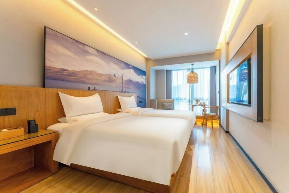 A hotel room with a large bed, bedside tables, a painting on the wall, and a view of the city.